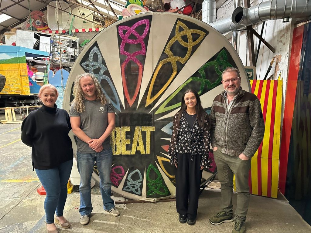 Four people smile for a photograph standing in front of a large drum decorated with celtic symbols and the words 'Beat'
