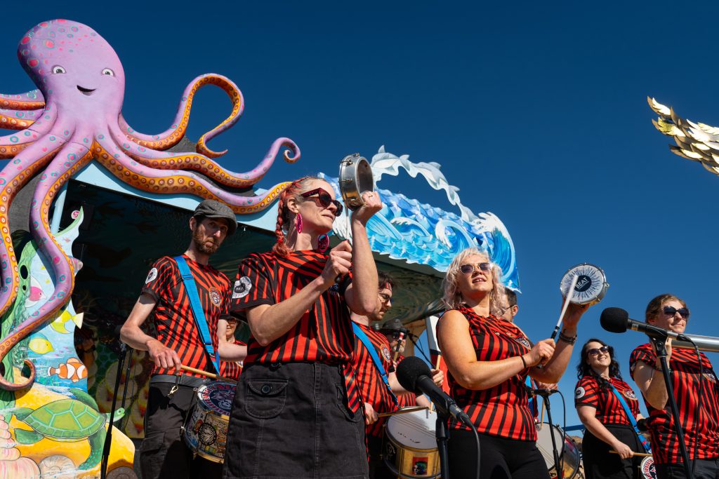 Image description: Six performers wearing black and red striped t-shirts are playing music outdoors using various percussion instruments