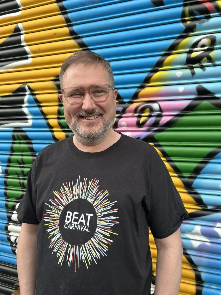 A man smiles for the camera. He is wearing glasses and a black t-shirt with the Beat Carnival logo. He stands in front of a colourful exterior building shutter
